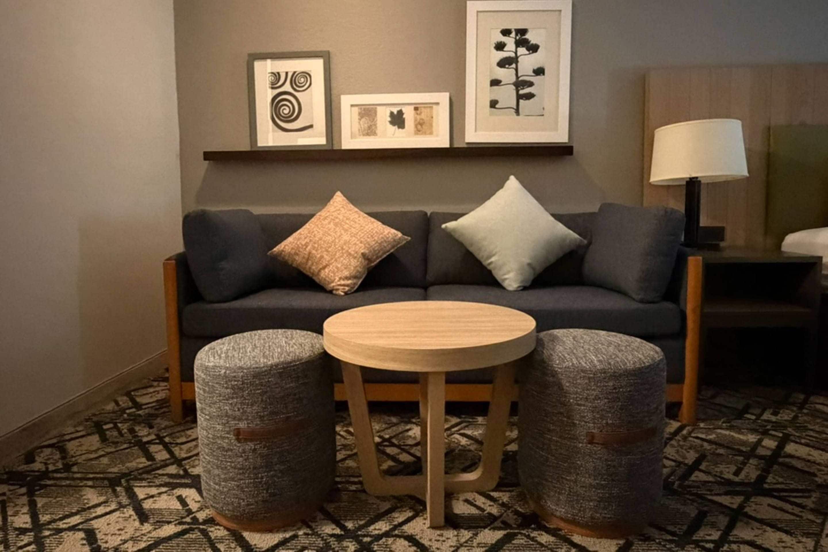 A modern living room with a dark grey sofa, two round wooden tables, and two grey woven ottomans. There are framed artworks on the wall above the sofa.