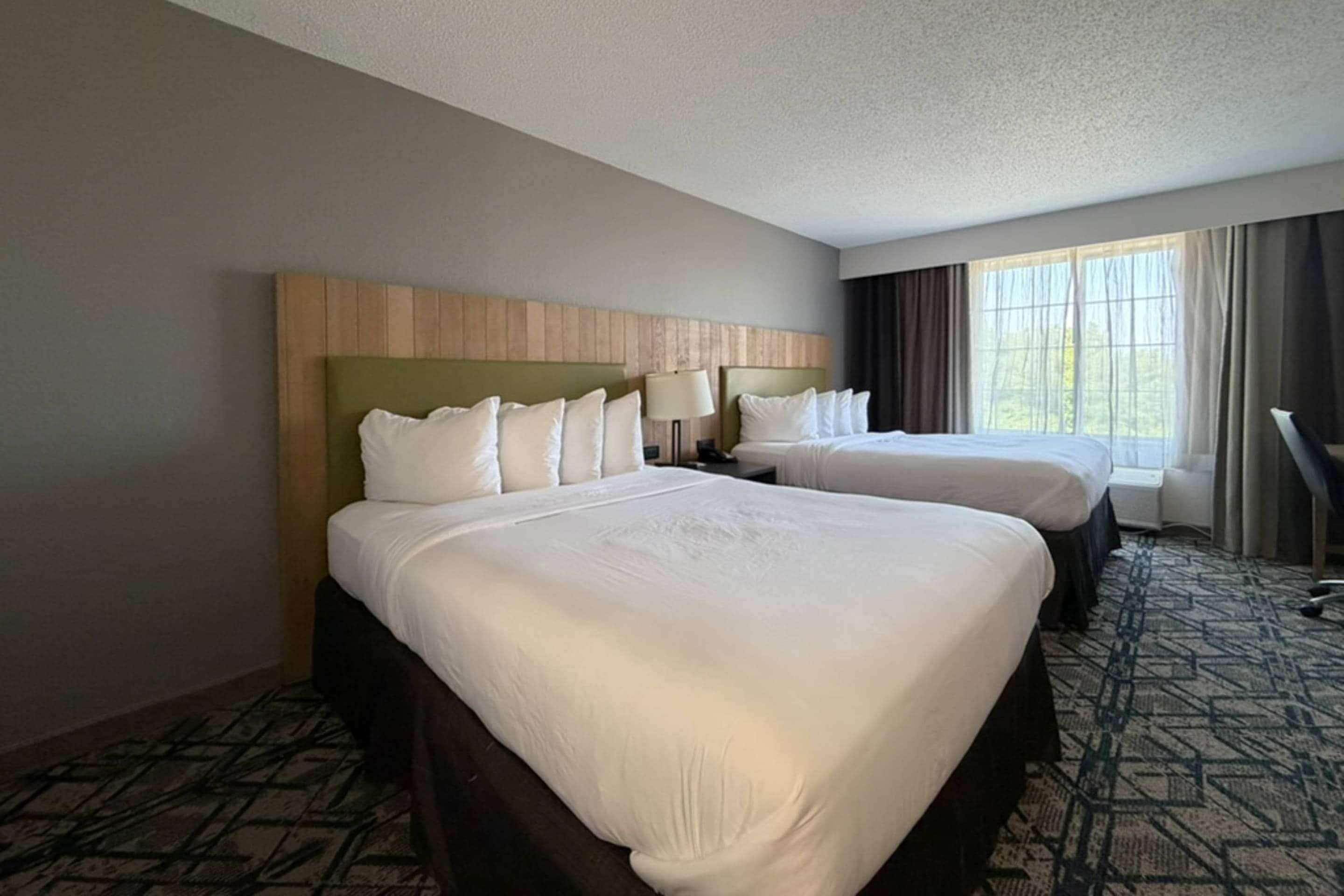 Habitación de hotel con dos camas, cabecera, mesita de noche, lámpara y ventana con cortinas.