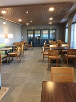 Uma sala de jantar ampla, com mesas e cadeiras de madeira, piso ladrilhado e teto com ripas de madeira e iluminação embutida.