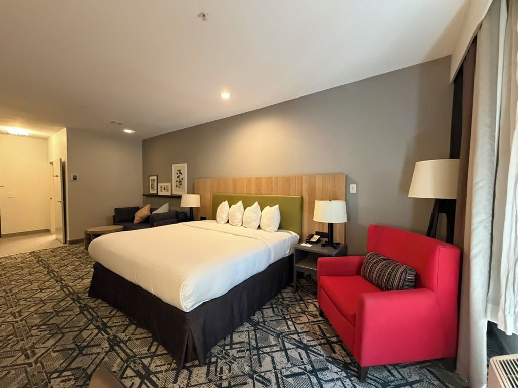 Habitación de hotel con una cama grande, un sillón rojo, una mesita de noche con una lámpara y un cabecero con luz.