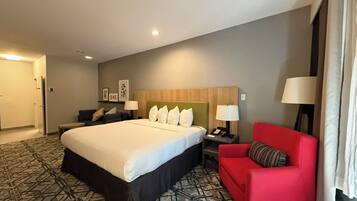 A hotel room with a large bed, a red armchair, a nightstand with a lamp, and a headboard with a light fixture.