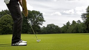 A person in a red and blue striped shirt, black pants, and white sneakers with black stripes, preparing to put a golf ball on a green.