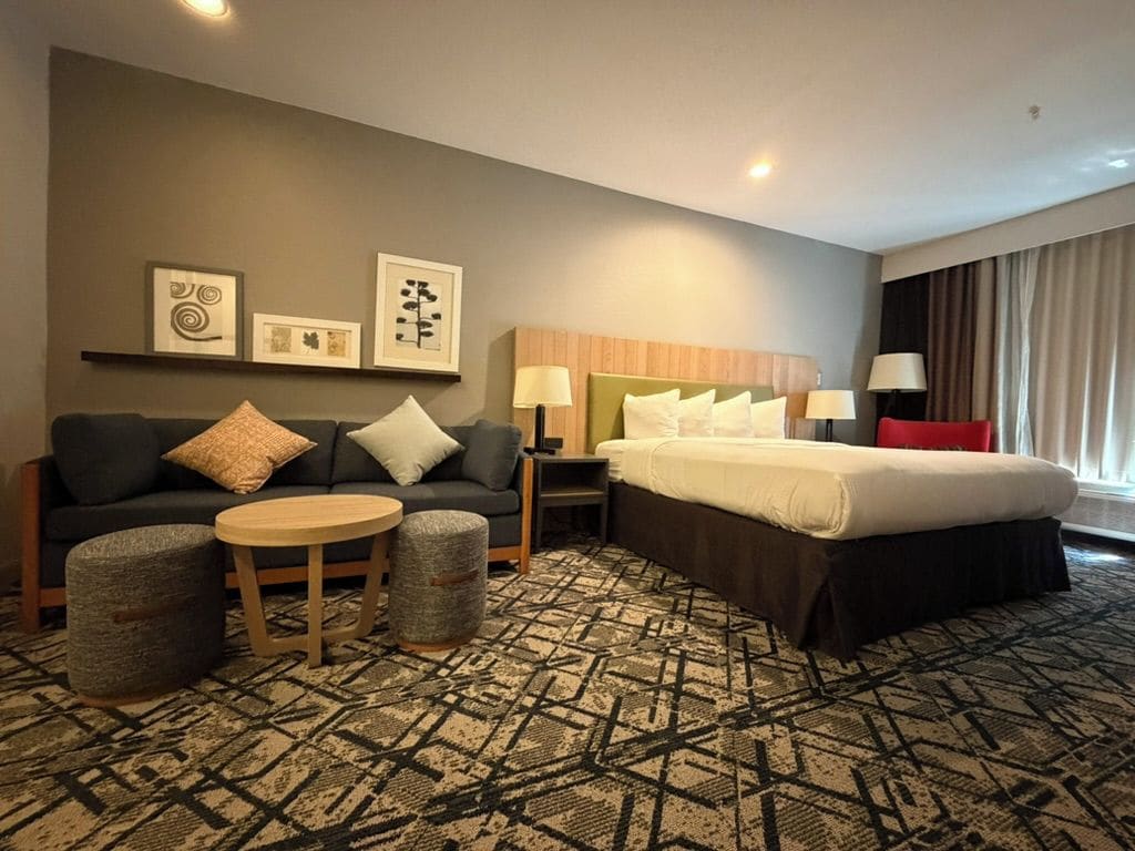 Quarto de hotel com cama, sofá, mesa pequena e cadeira. Há luminárias fixadas na parede e uma janela com cortinas.