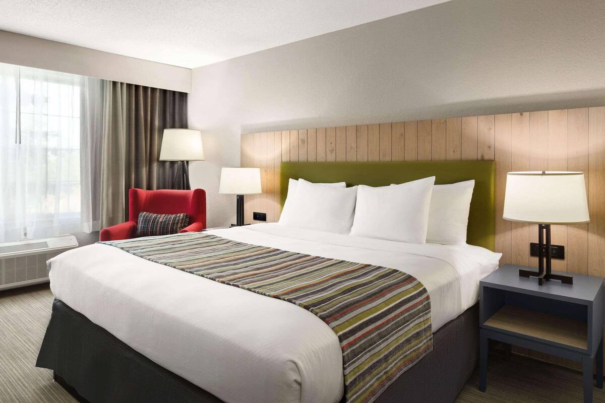 A hotel room with a large bed, two bedside tables with lamps, a red armchair, and a view of the outdoors through a window with curtains.