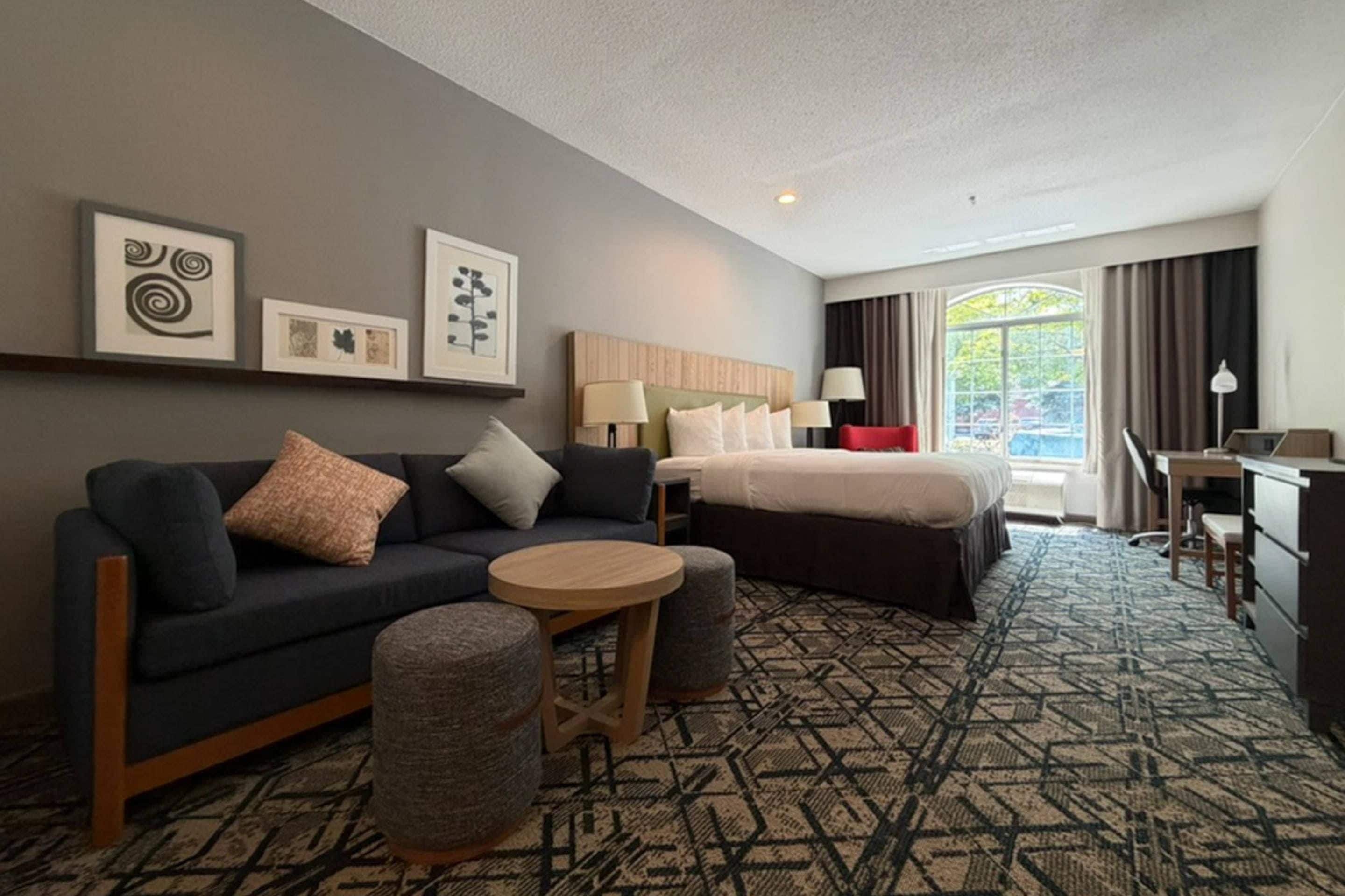 Une chambre d’hôtel avec un canapé de couleur sombre, une table ronde en bois, un lit avec une literie blanche, un bureau avec une chaise et une grande fenêtre avec des rideaux.