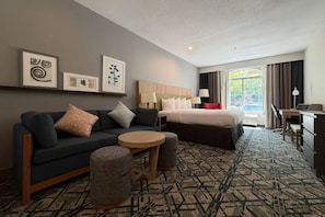 A hotel room with a dark-colored sofa, a round wooden table, a bed with white bedding, a desk with a chair, and a large window with curtains.