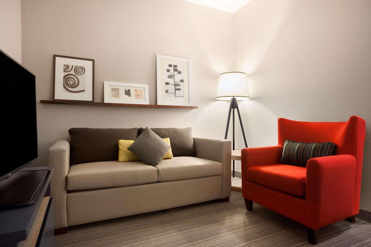 A living room with a beige sofa, a red armchair, and a floor lamp. There are framed pictures on the wall.
