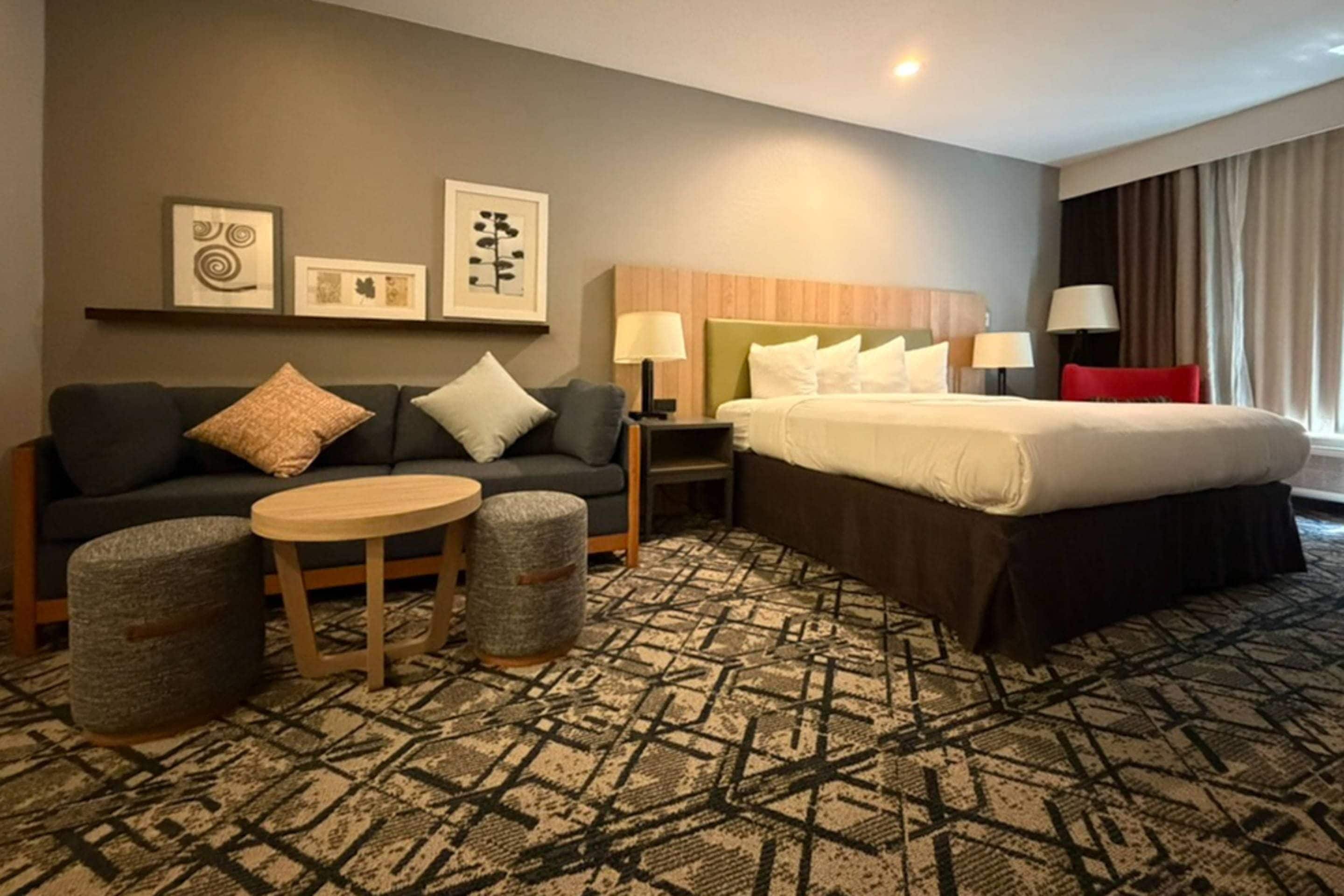 Une chambre d’hôtel comprenant un lit, un canapé, une petite table, deux poufs, une table de chevet et des œuvres d’art murales.