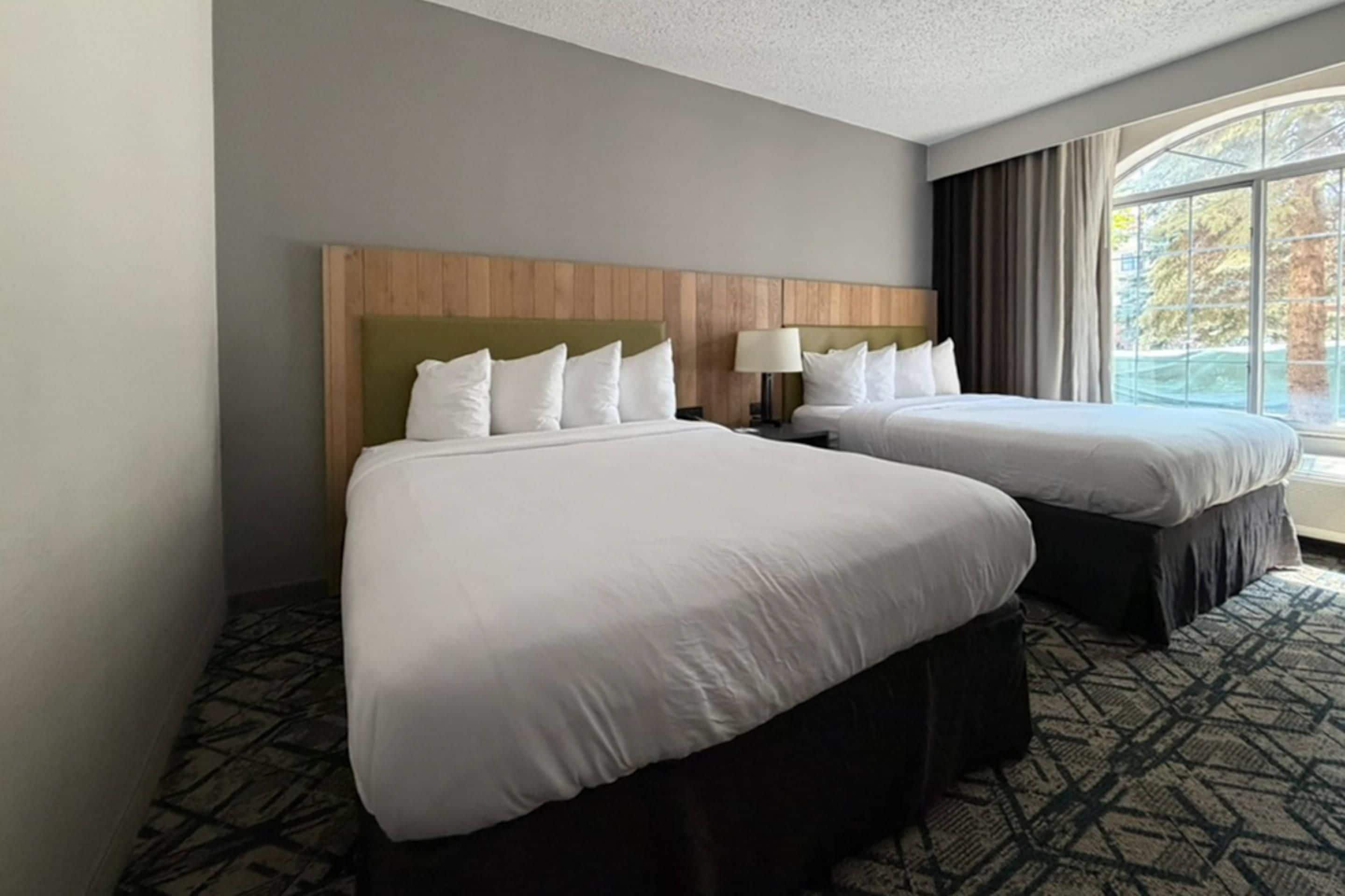 A hotel room with two beds, a wooden headboard, a nightstand with a lamp, and a large window with curtains.