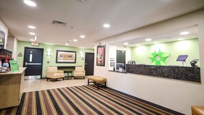 Zona de recepción de hotel con alfombra a rayas, mostrador de recepción, un cartel de 'Welcome', un logotipo verde y un cartel que dice 'Buenos días'.