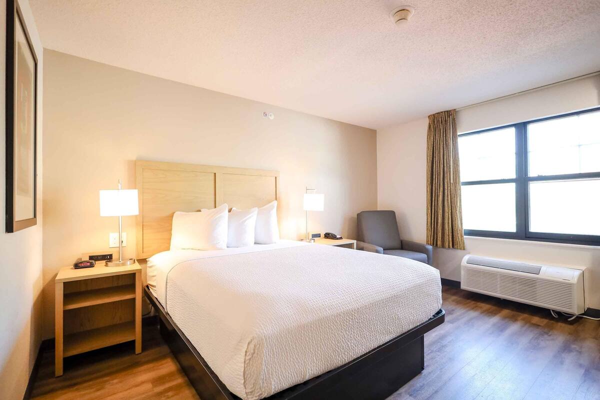 Ein Hotelzimmer mit einem großen Bett, zwei Nachttischen mit Lampen, einem Sessel, einem Fenster mit Vorhängen und einer Wand-Klimaanlage.