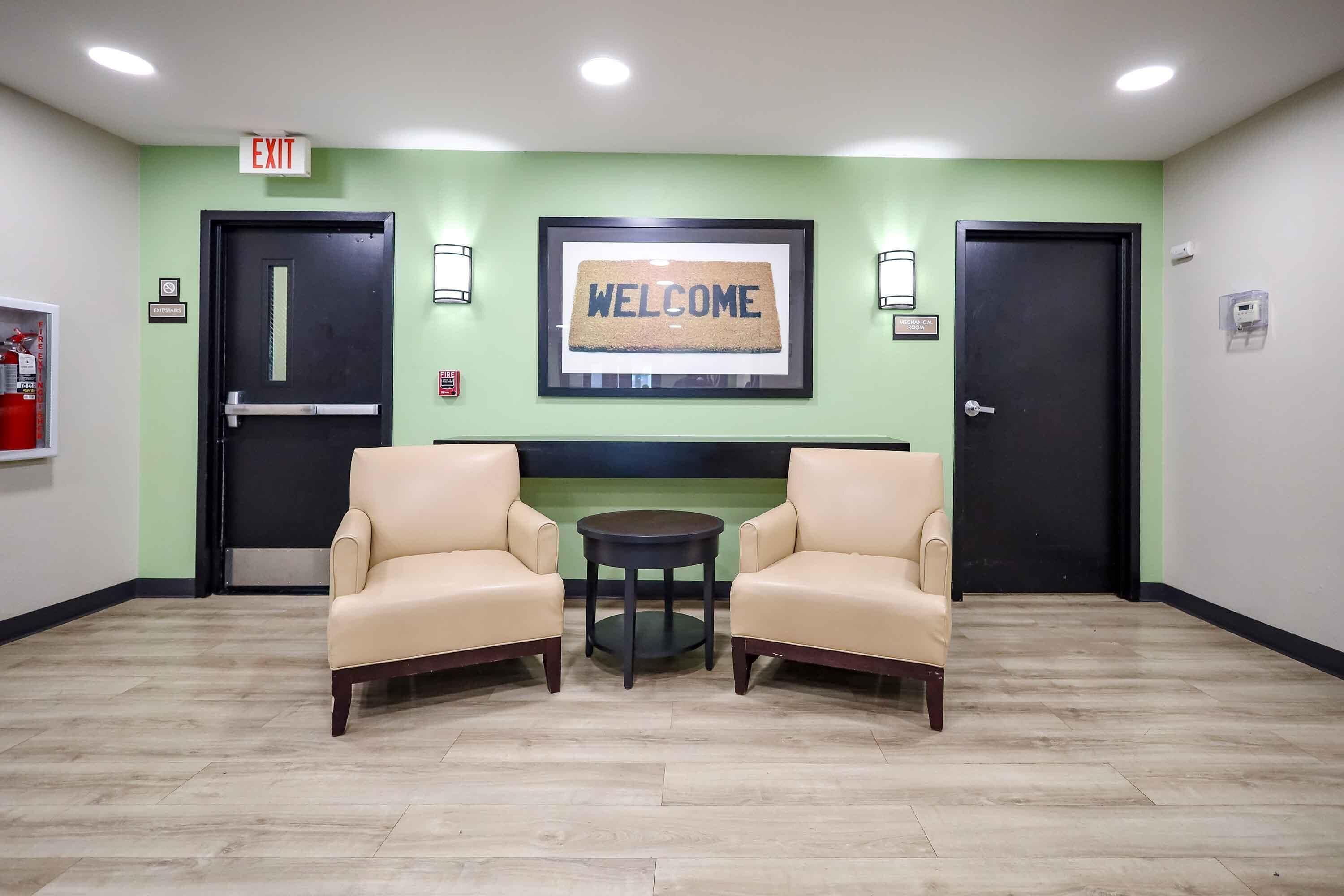 A lobby with two beige armchairs, a round table, a welcome sign, and two black doors.