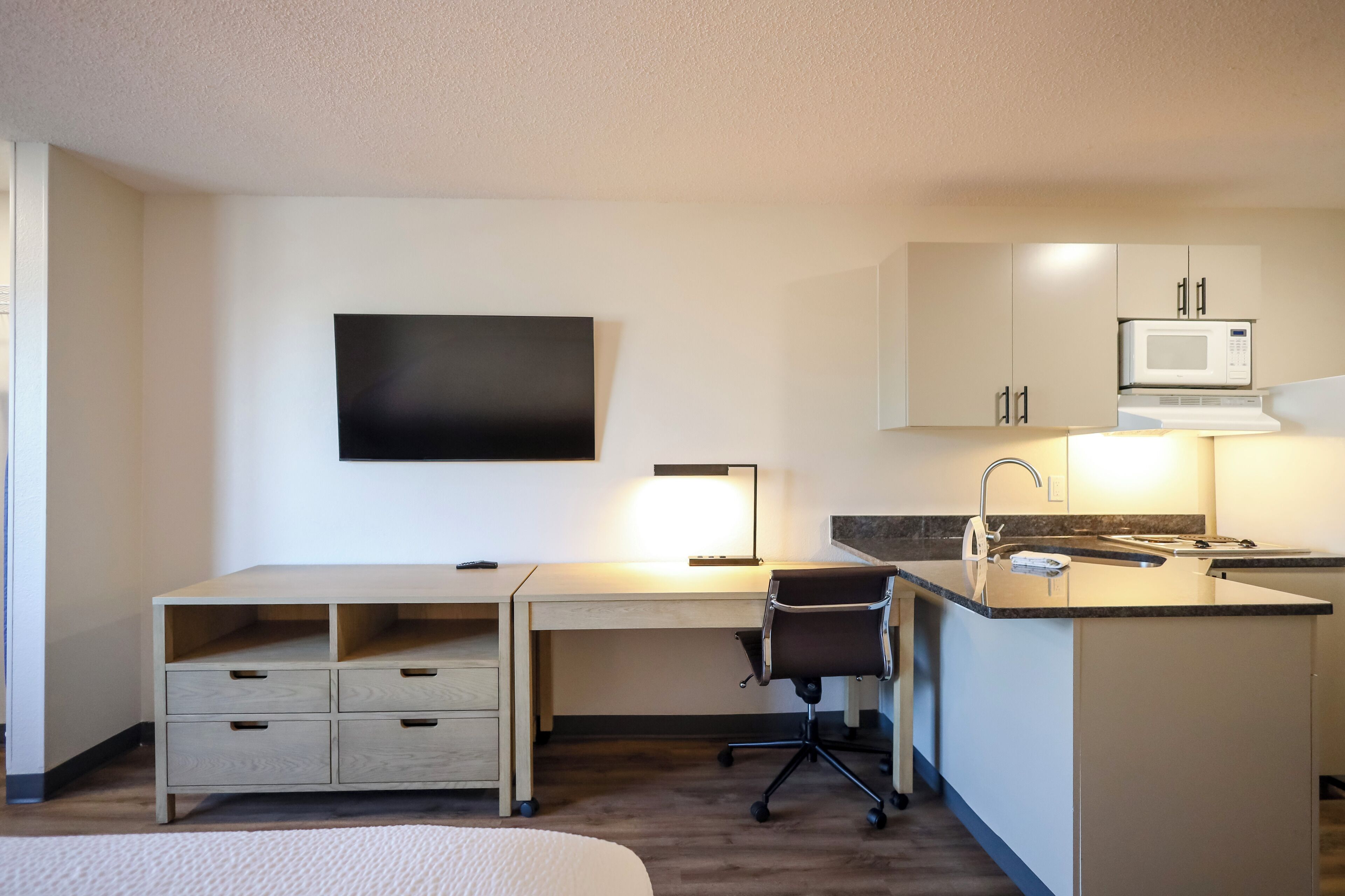 A hotel room with a bed, a desk with drawers, a chair, a TV mounted on the wall, a microwave, and a small kitchen area with a sink.