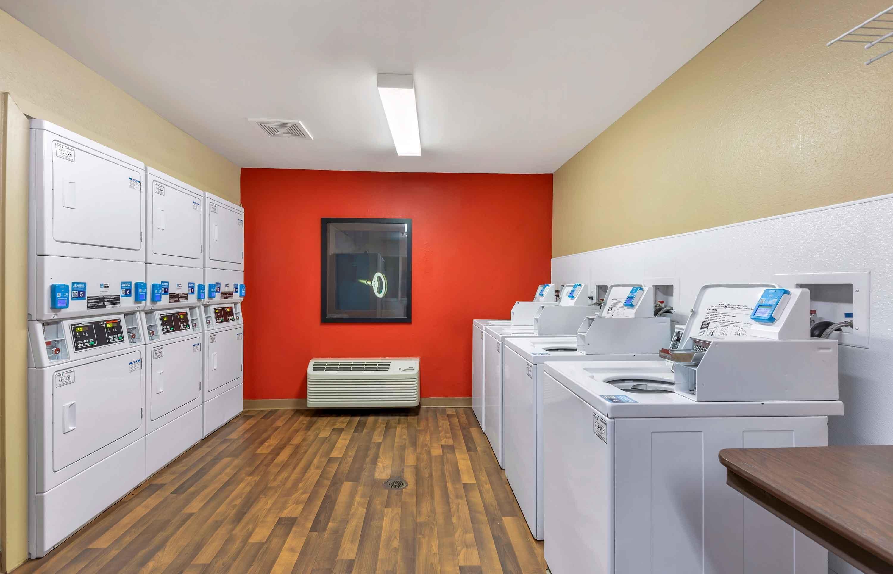 A laundry facility with multiple washing machines and dryers, a red wall, and a vent on the floor.