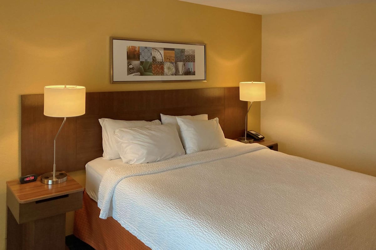 A neatly made bed with white linens, two bedside lamps, a clock, and a framed artwork above the bed.