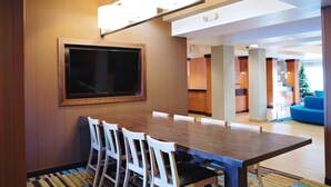 Uma área de refeições moderna com uma mesa de madeira e cadeiras brancas, uma grande TV de tela plana na parede e um lustre de luz clara.