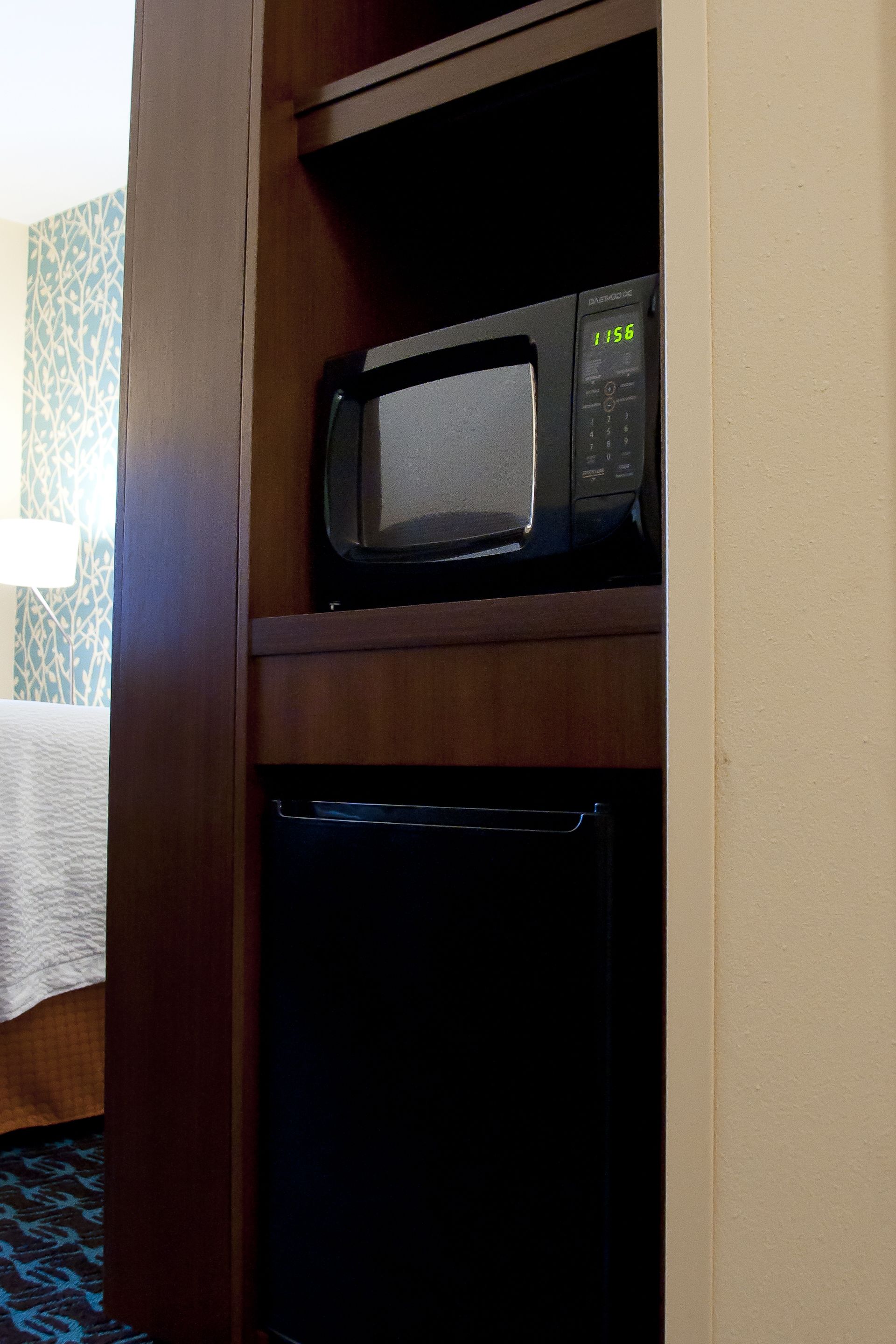 A microwave on a wooden shelf above a black appliance, likely a dishwasher, in a hotel room with patterned wallpaper and a bed with a white bedspread.