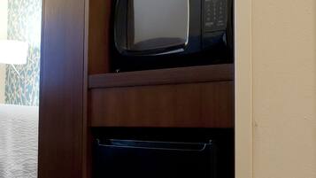 A microwave on a wooden shelf above a black appliance, likely a dishwasher, in a hotel room with patterned wallpaper and a bed with a white bedspread.