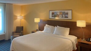 Quarto de hotel com uma cama grande, duas luminárias de cabeceira, uma cadeira, uma mesinha de cabeceira e um quadro na parede.