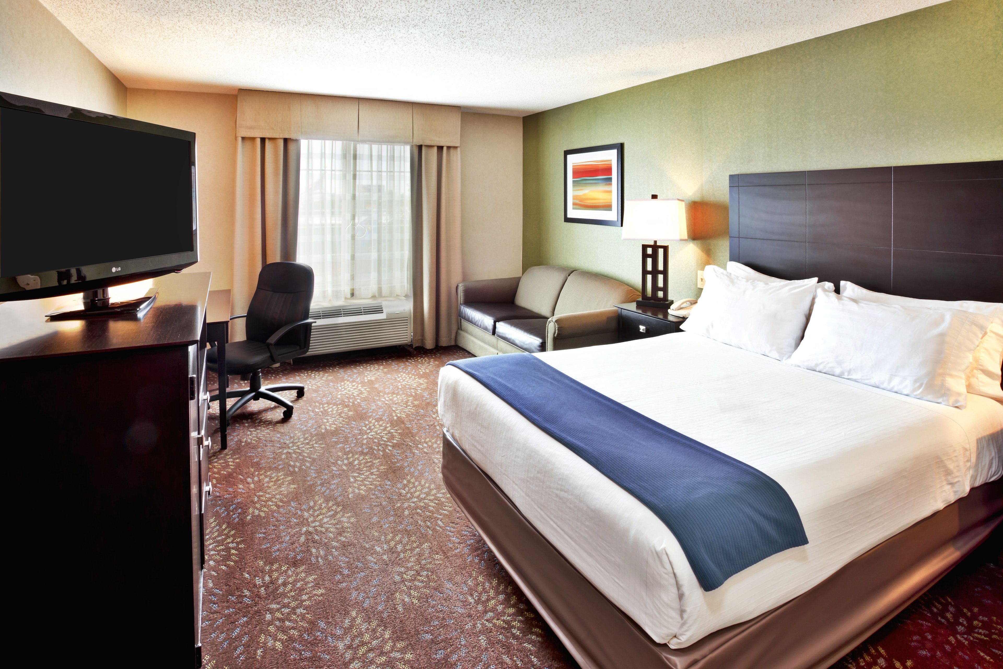 A hotel room with a large bed, a desk with a TV, a chair, a sofa, and a wall-mounted lamp.