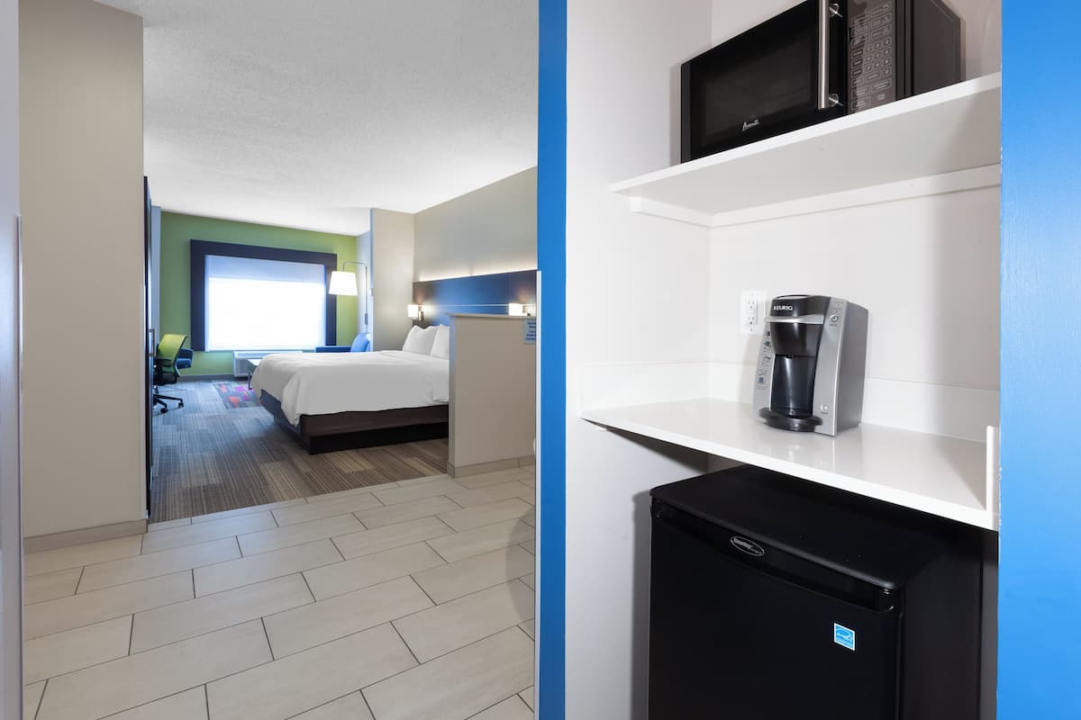 A hotel room with a bed, desk, chair, and a kitchenette area with a coffee maker and refrigerator.