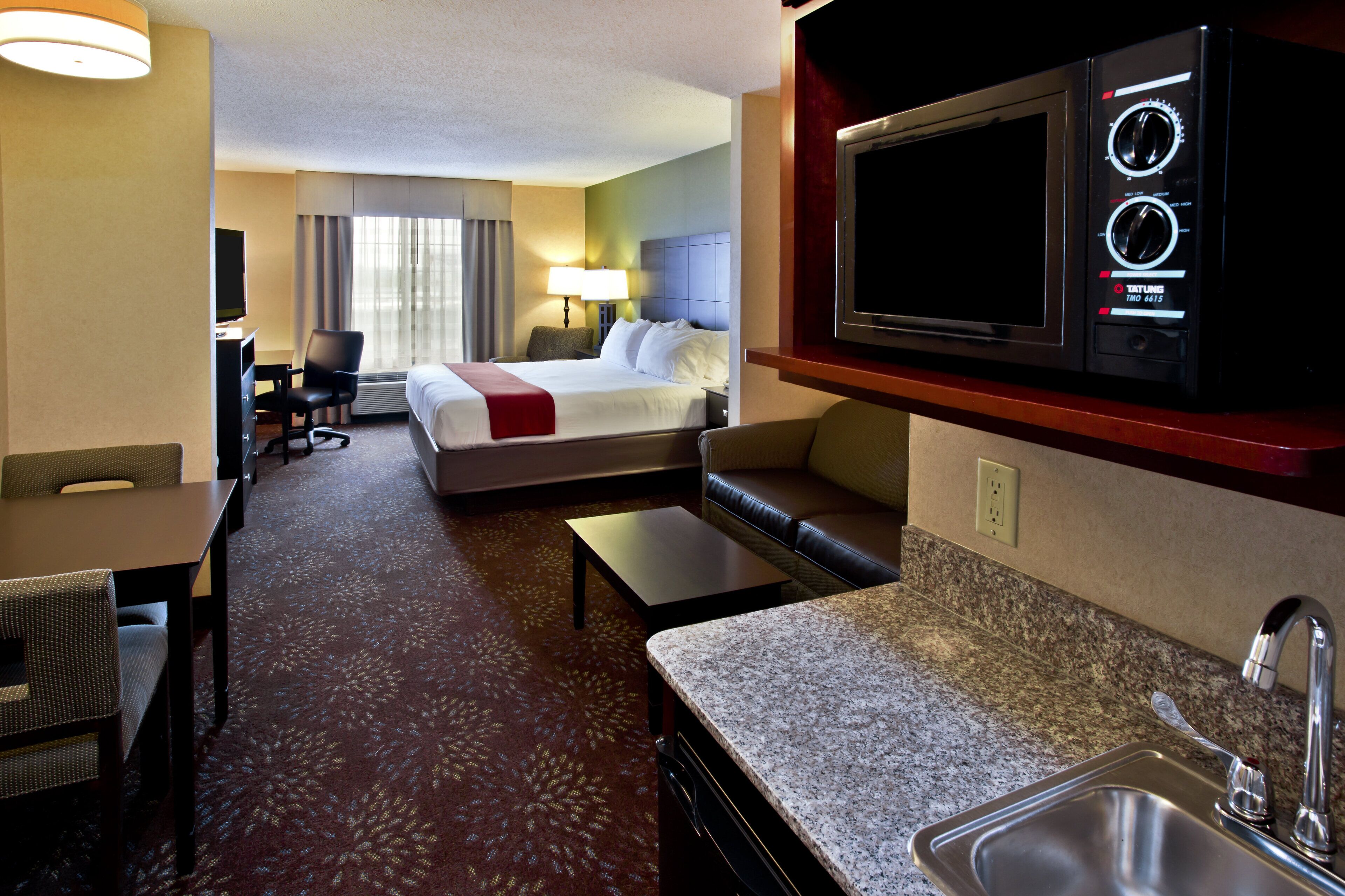 A hotel room with a bed, a desk, a chair, a sofa, a television, a microwave, and a sink.