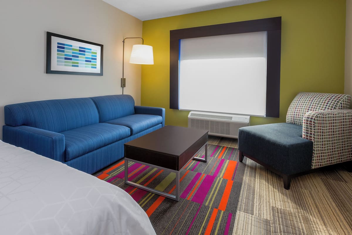A hotel room with a blue sofa, a small table, a patterned armchair, a large window with blinds, a framed picture on the wall, and a colorful striped carpet.