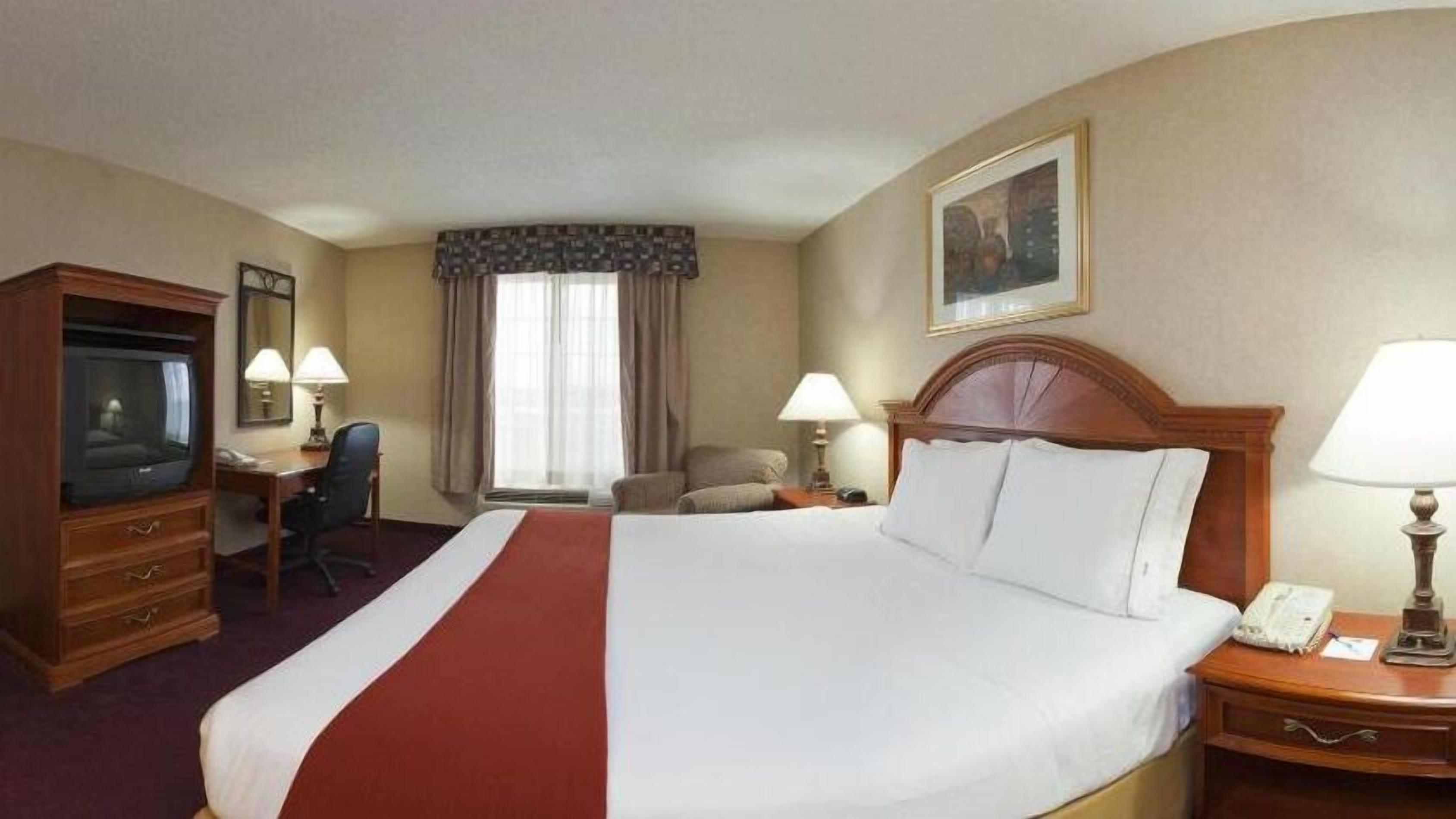 A hotel room with a large bed, a desk with a chair, a television, a lamp, and a painting on the wall.