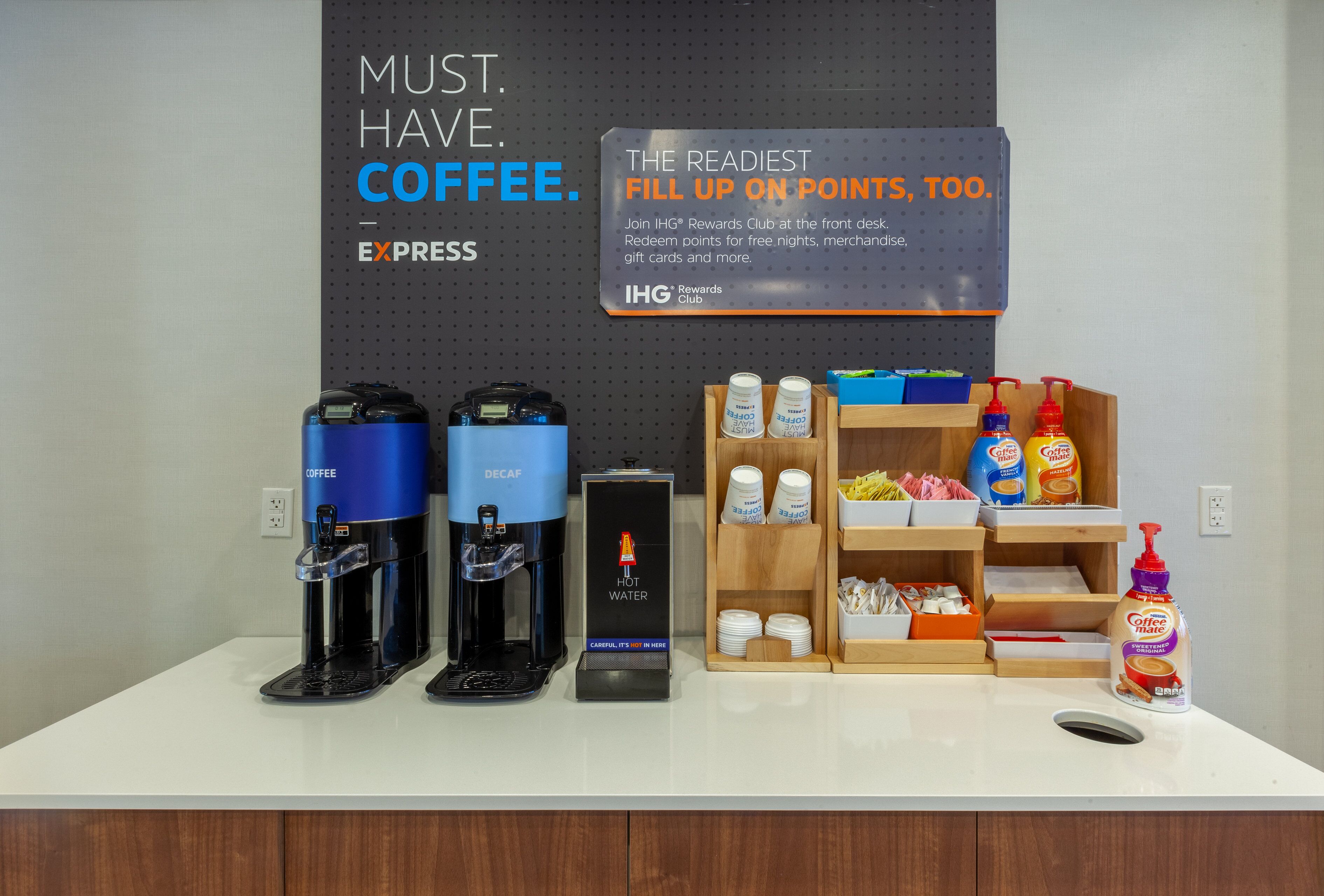 A coffee station with two coffee makers, a sign promoting the IHG Rewards Club, and a display of coffee products and condiments.