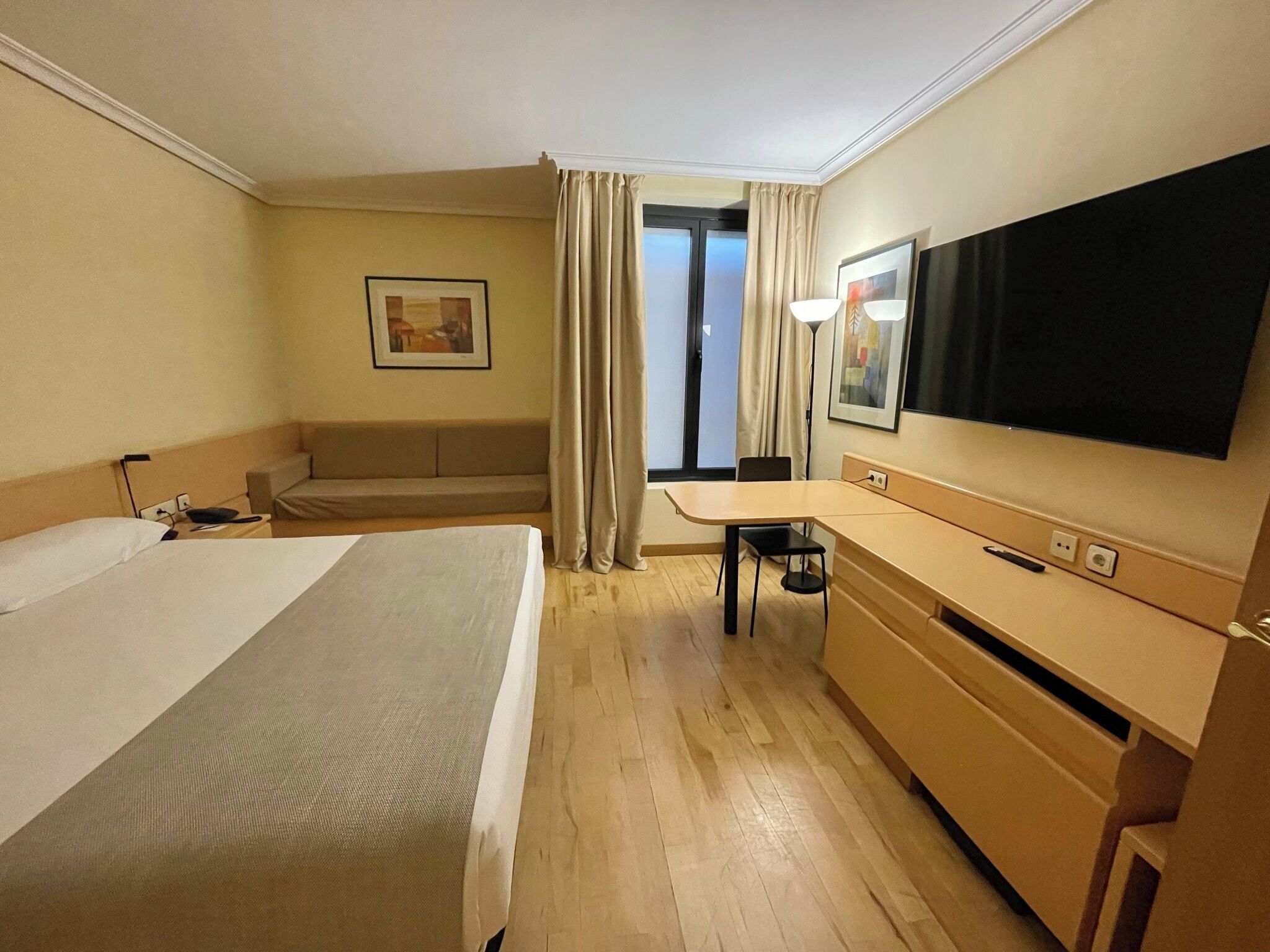 Habitació d'hotel amb llit, escriptori, cadira, televisió i finestra amb cortines.