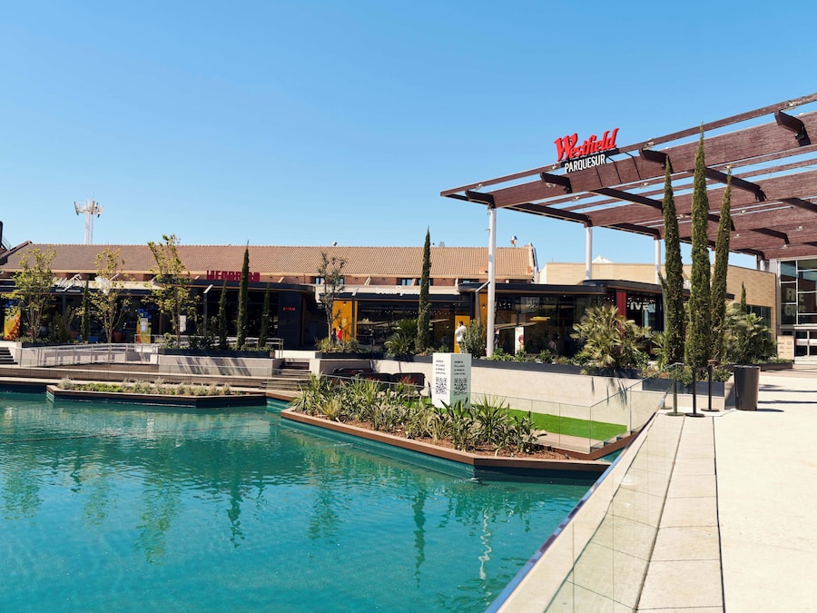 Uma área de piscina com piscina azul límpida, o edifício Westfield Parquesur e uma galeria coberta com assentos.