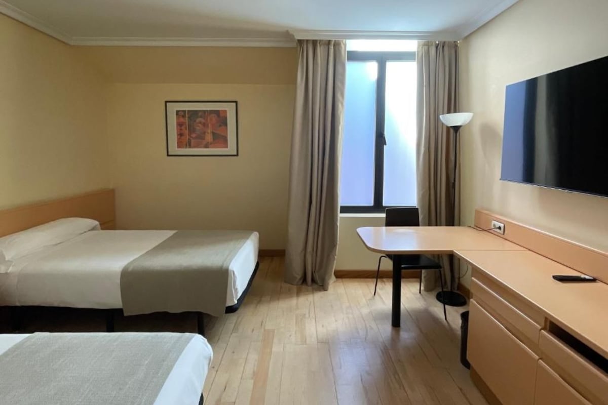 A hotel room with two beds, a desk, a TV, and a window with curtains.