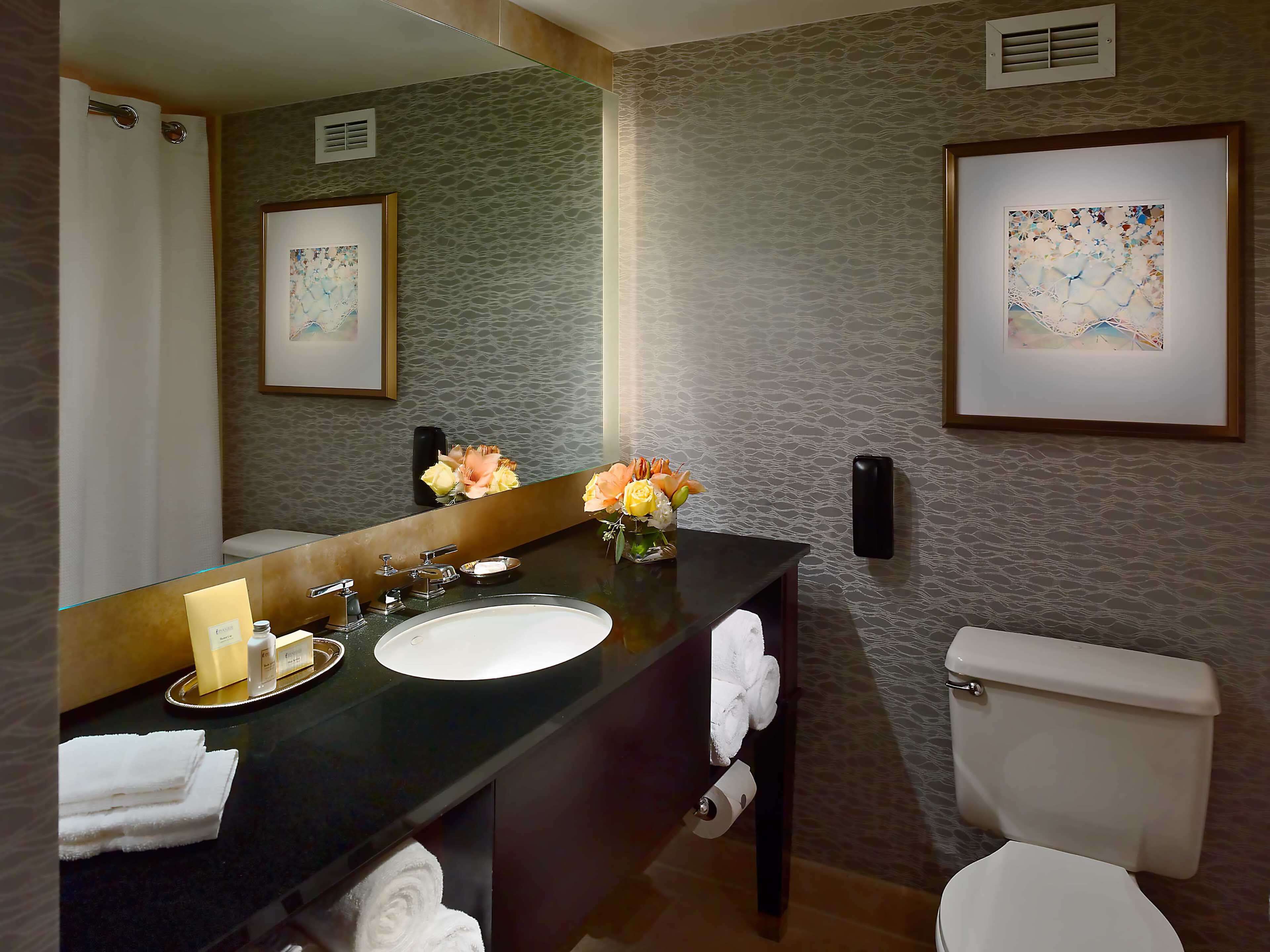 A bathroom with a large mirror, a sink, a toilet, and two framed artworks on the walls.