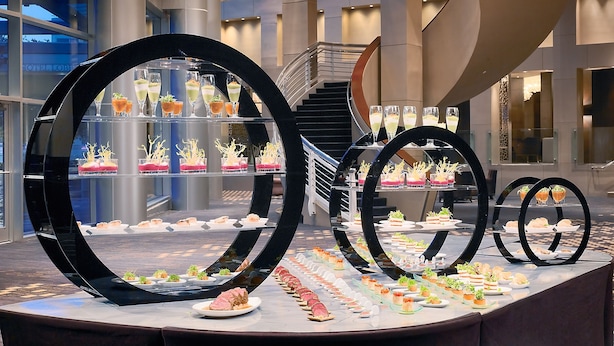 A modern hotel interior with a central spiral staircase, large glass windows, and a buffet area with various food items.