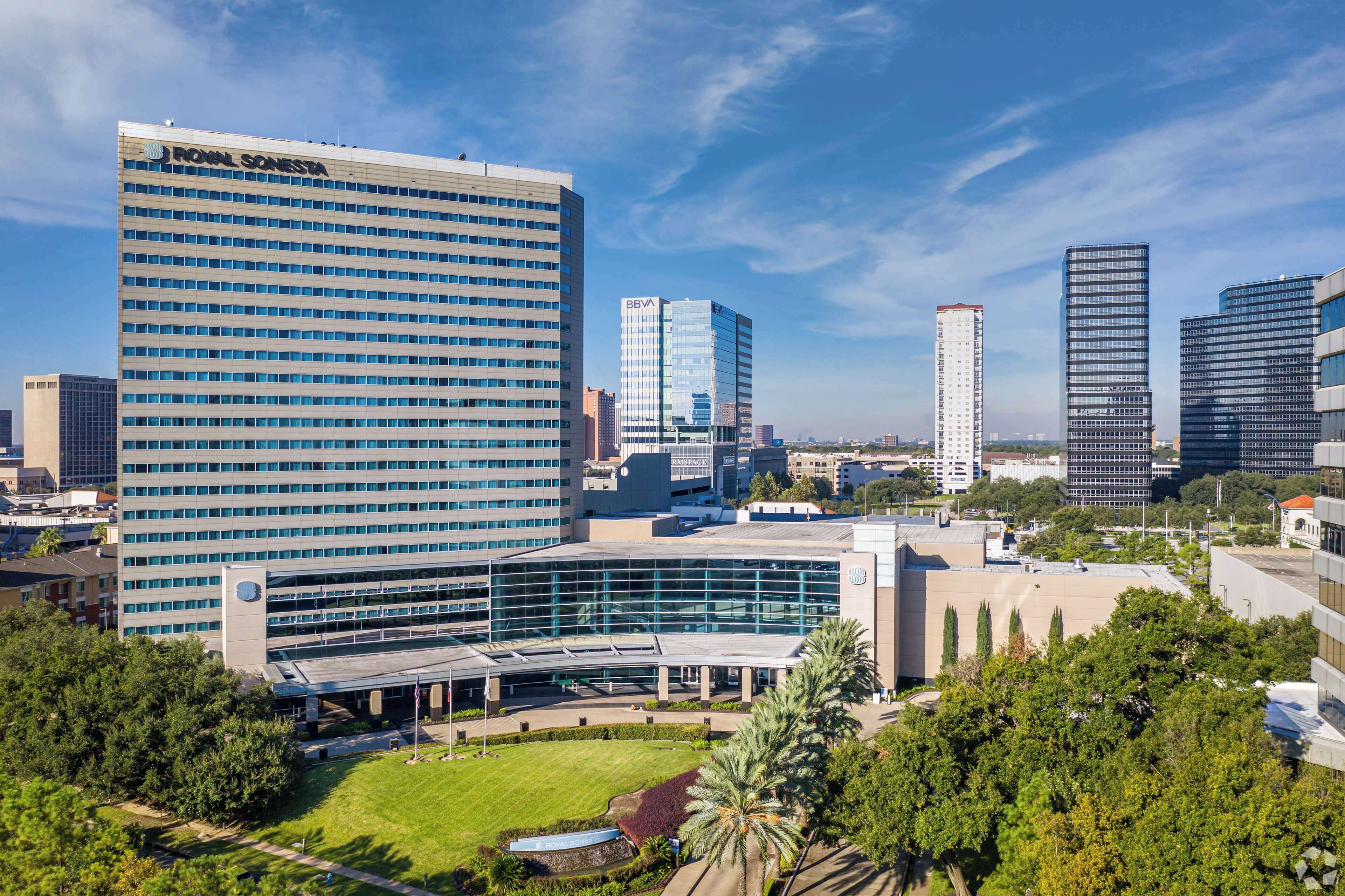 A high-rise building with a glass facade, a large parking structure, and surrounding greenery.