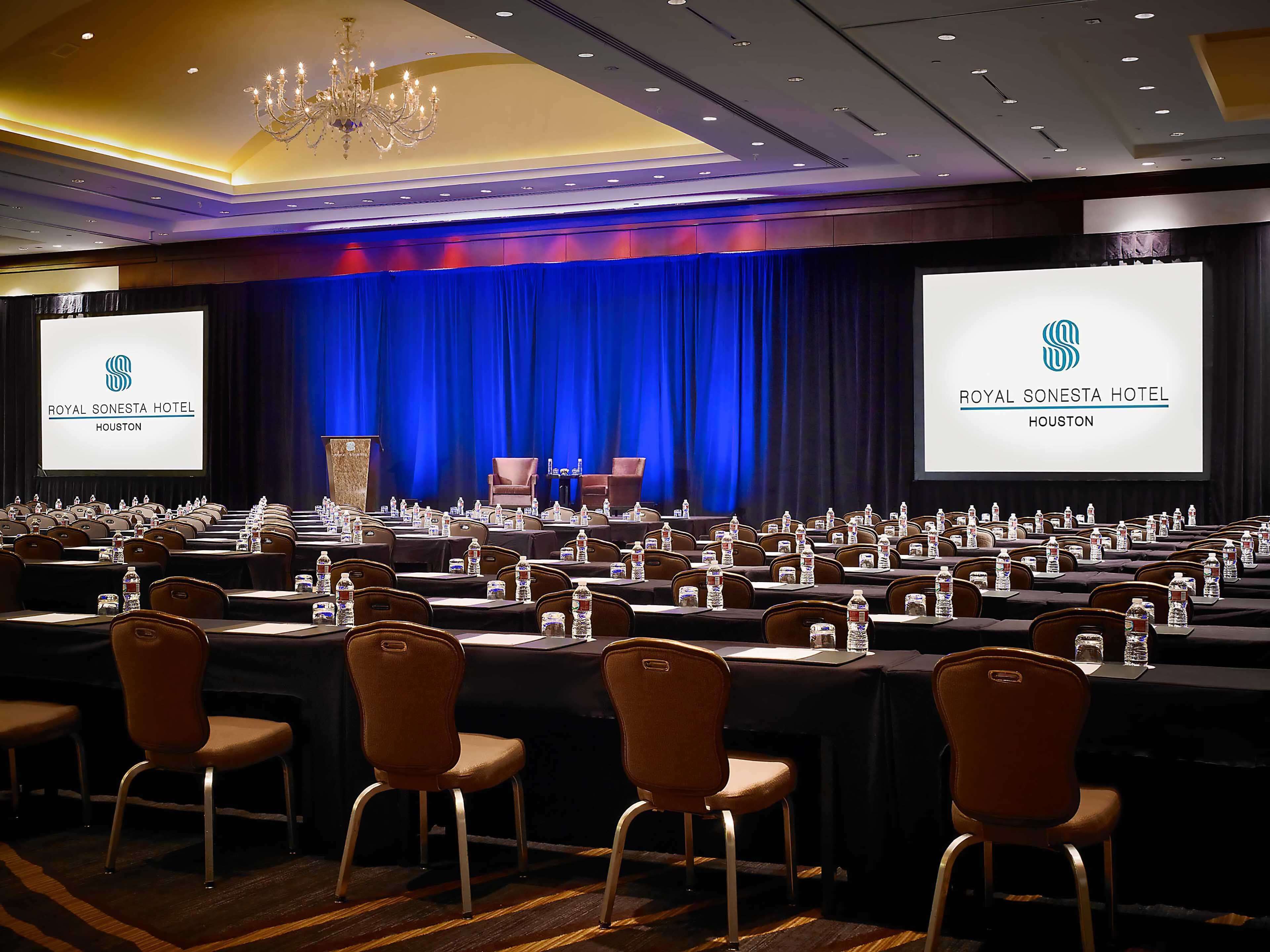 A conference room at the Royal Sonesta Hotel in Houston with rows of tables, chairs, and a stage with a blue curtain backdrop.