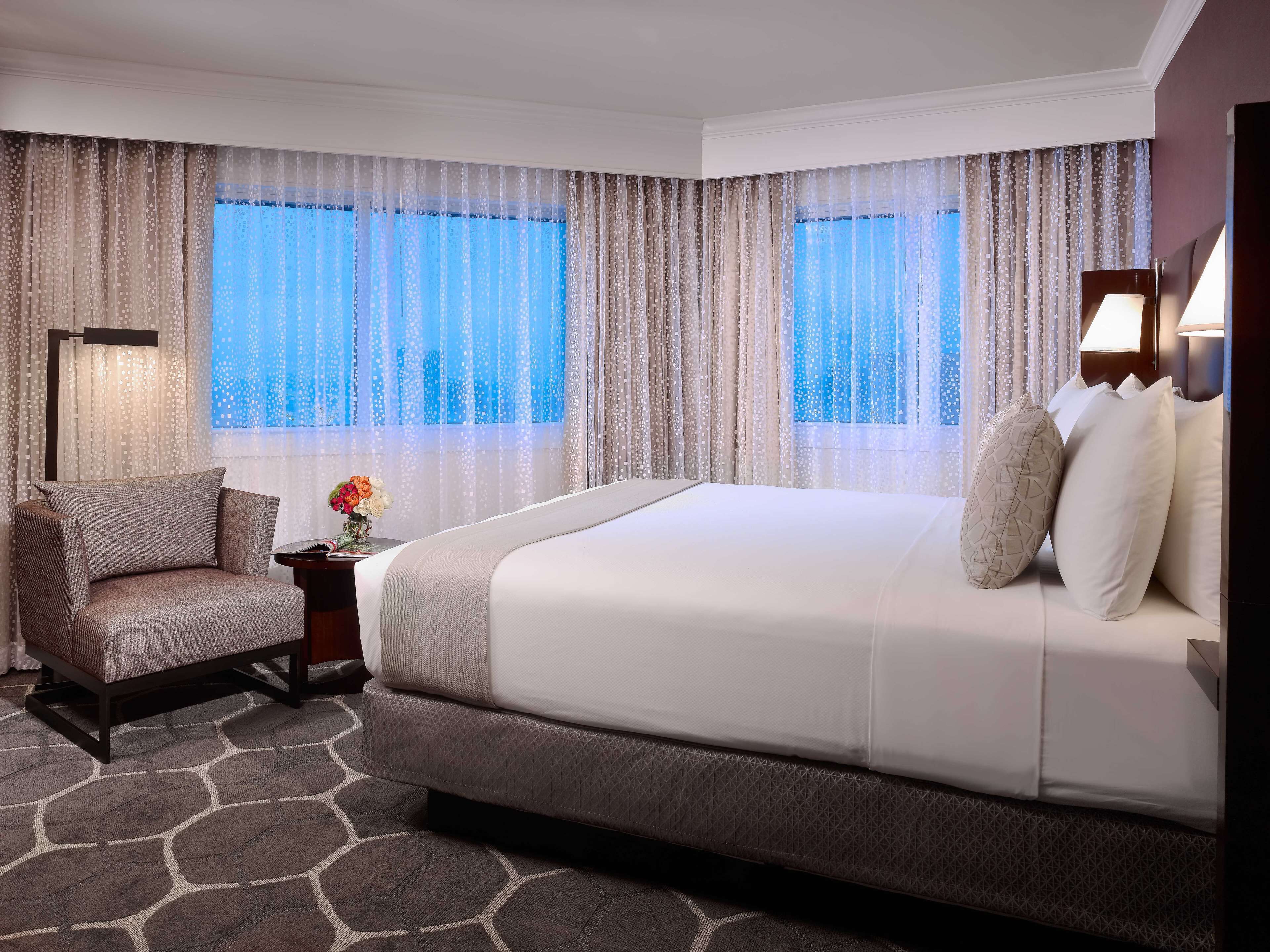 A hotel room with a large bed, a chair, a nightstand with a lamp, and a view of the city through sheer curtains.
