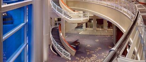 Interior de hotel com vários níveis, uma escadaria espiral central, elevador de vidro e carpete estampado.