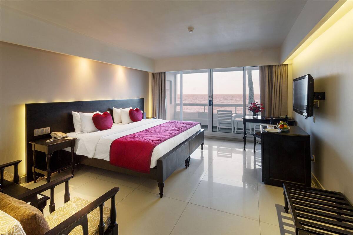 A hotel room with a large bed, a desk, a television, and a balcony with a view of the sea.