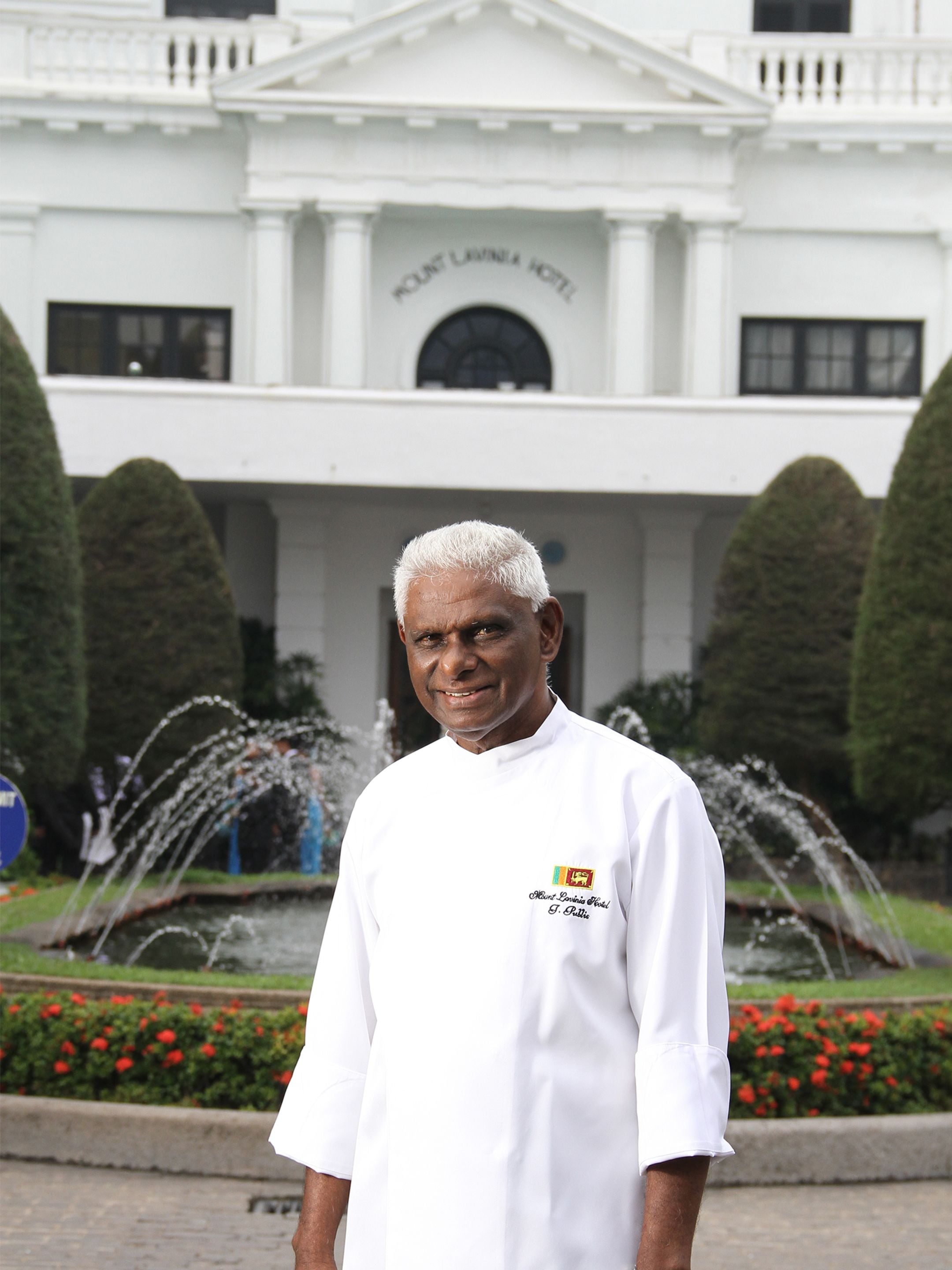 Ein Koch in Uniform steht vor einem Gebäude mit Springbrunnen und gepflegten Büschen.
