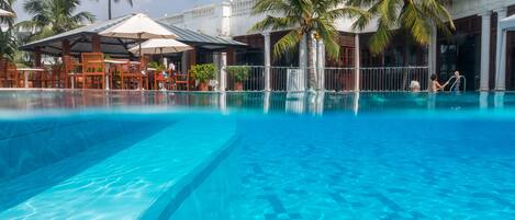 A large outdoor swimming pool with clear blue water, surrounded by palm trees and outdoor seating areas.