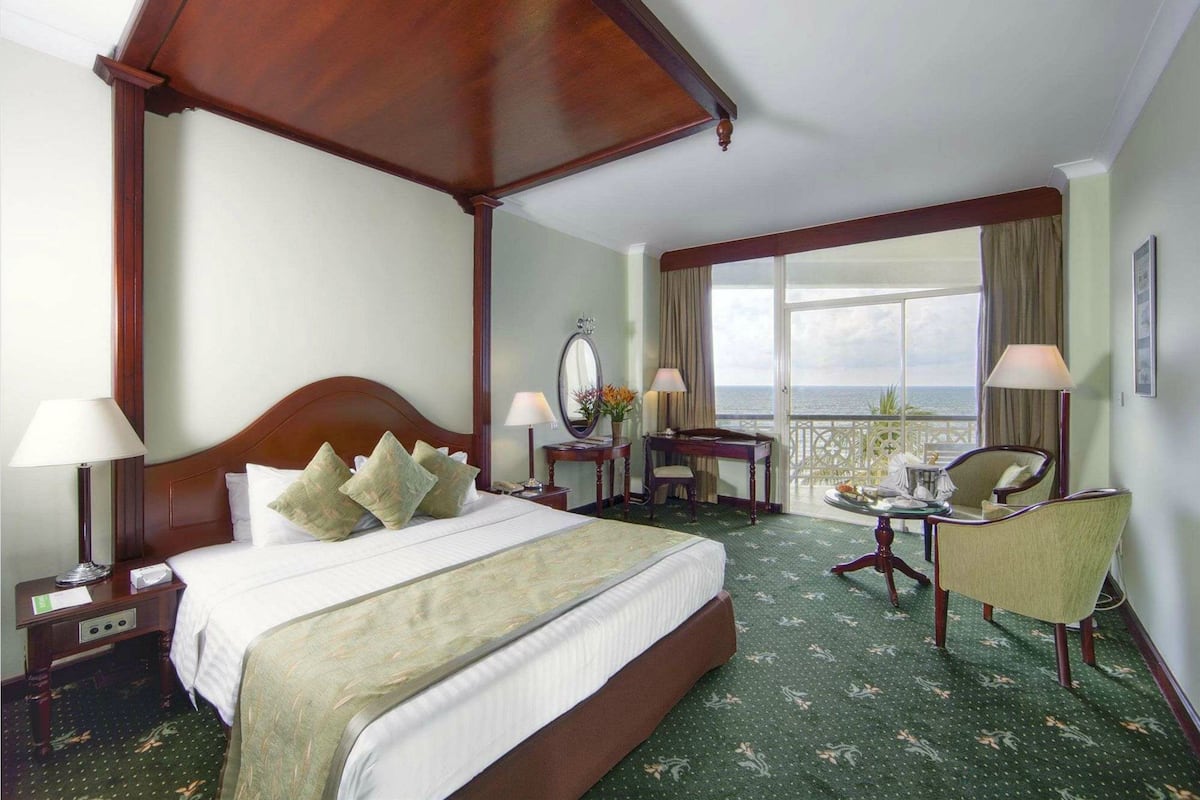 A hotel room with a four-poster bed, a desk with a chair, a balcony with a table and chairs, and a large window offering a view.