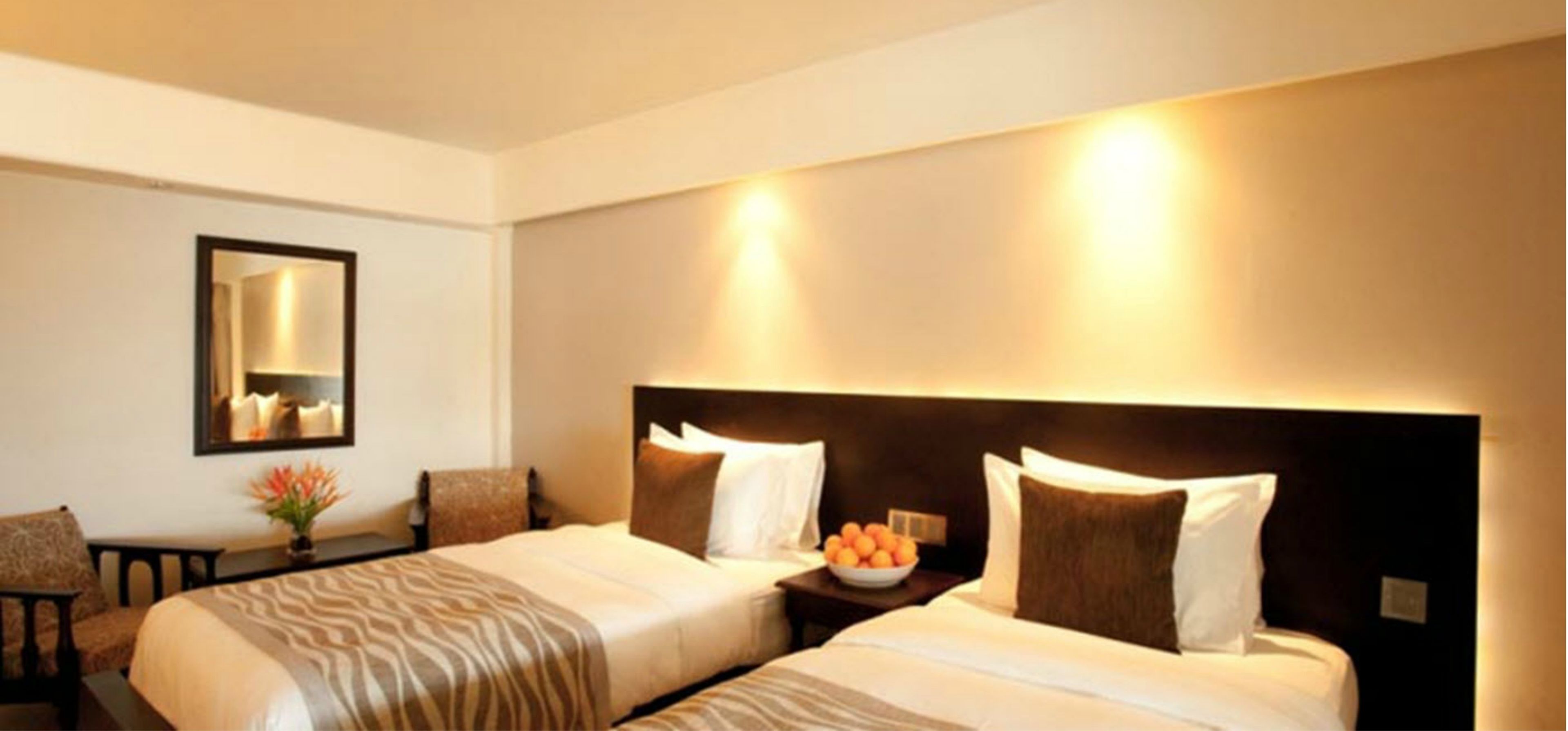 Une chambre d’hôtel avec deux lits, une table de chevet sur laquelle se trouve un bol de fruits, un miroir et une chaise.