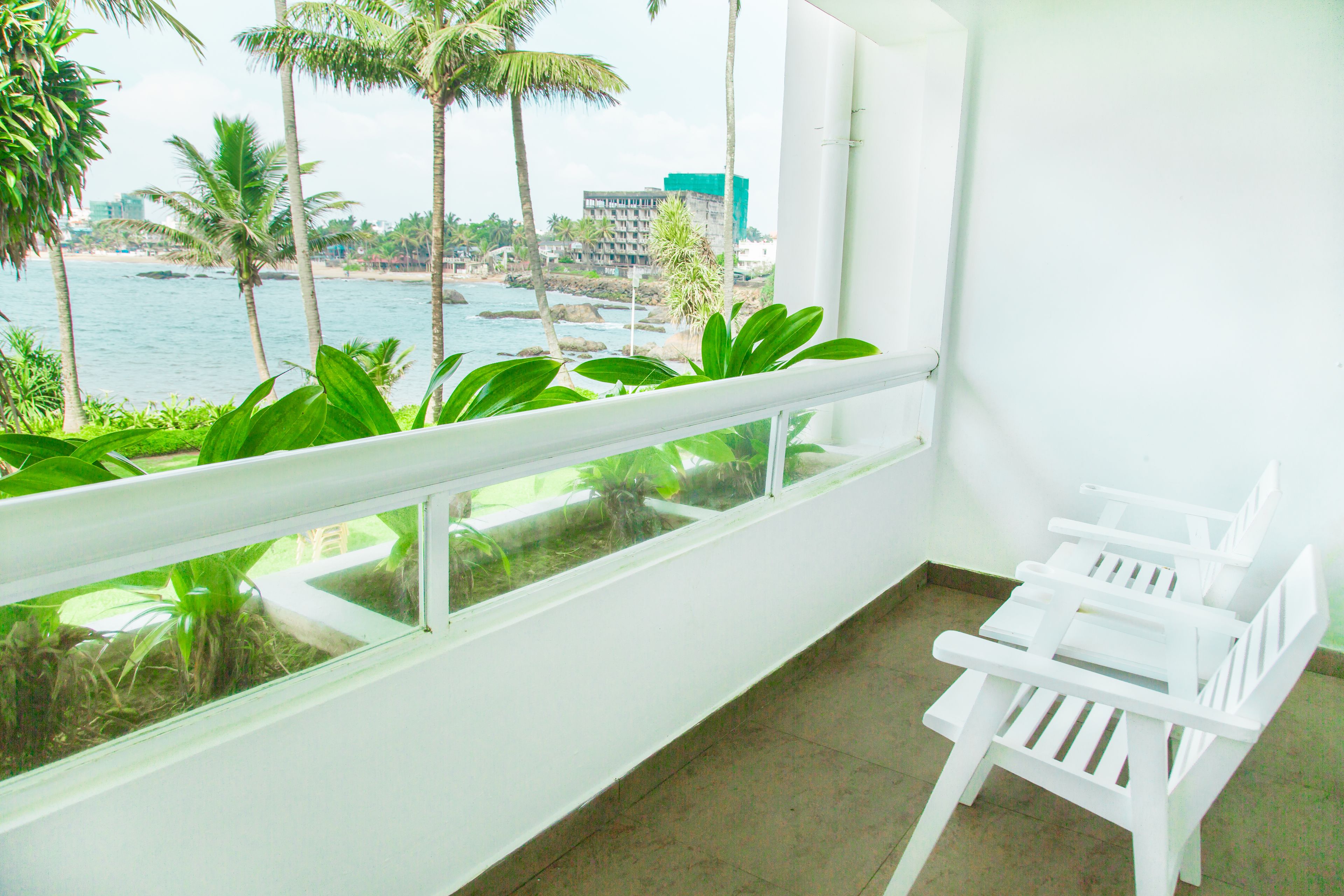 A balcony with a white railing, a white chair, and a view of the ocean and palm trees.