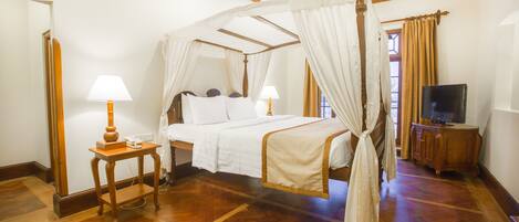 A four-poster canopy bed with a wooden frame and white bedding, a bedside table with a lamp, and a television on a wooden cabinet.