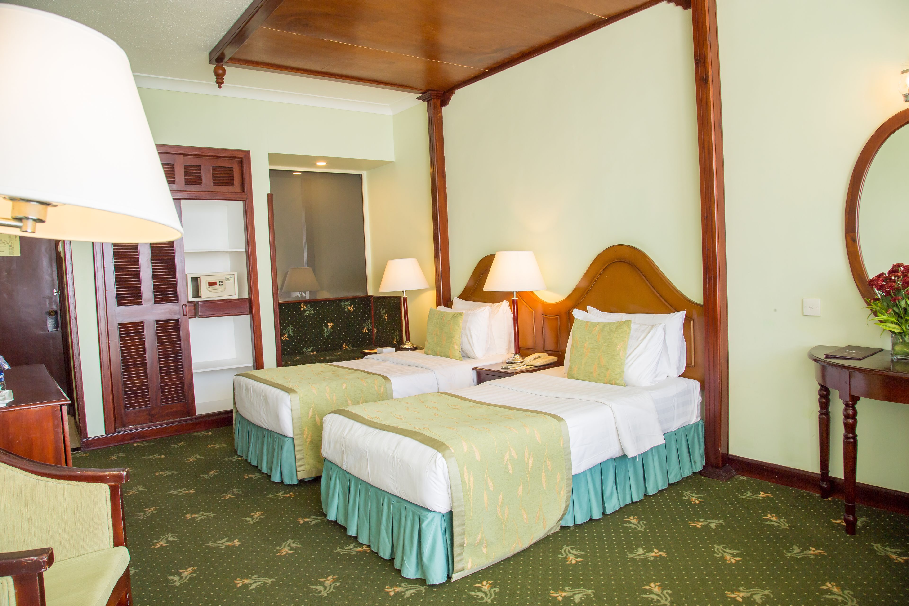 A hotel room with two beds, a wooden headboard, a desk with a lamp, a mirror, and a small table with a vase.