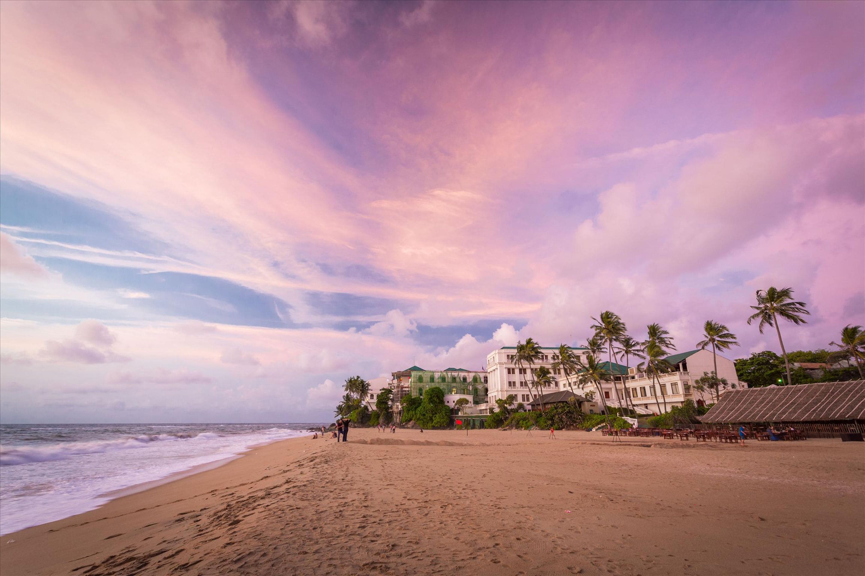 Una playa con un edificio al fondo, palmeras y cielo despejado.