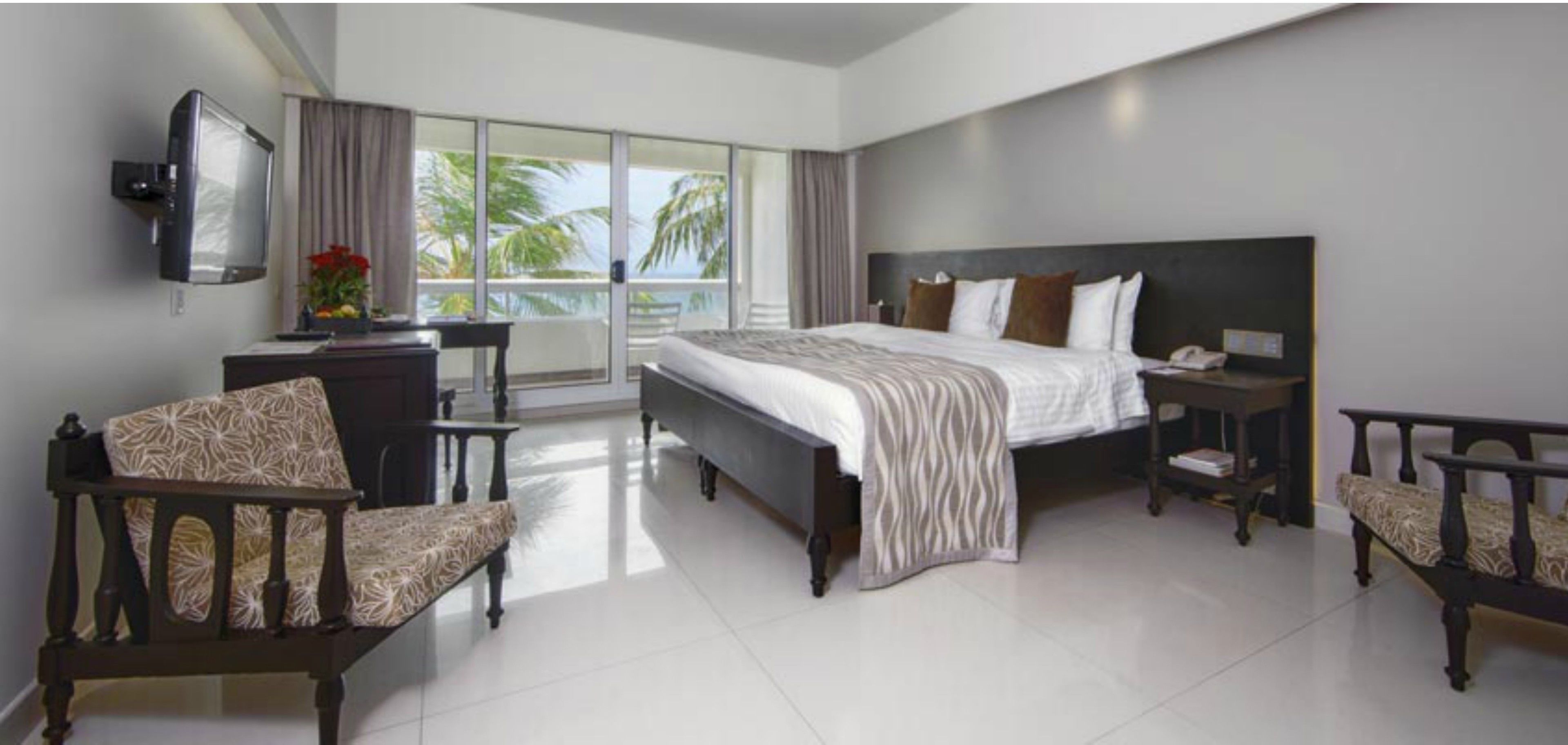 A hotel room with a bed, a desk, a chair, a TV, and a balcony with a view of palm trees.