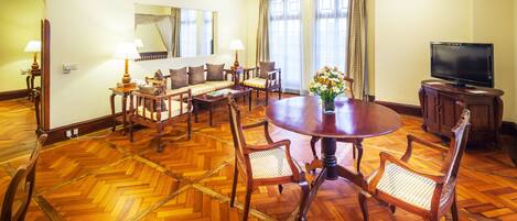 A spacious room with a wooden herringbone floor, a round dining table surrounded by chairs, a sofa set, a television, and a ceiling fan.