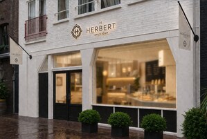 En butiksfacade med et skilt der siger 'DE HERBERT AMSTERDAM' og et stort vindue, der viser indretningsbelysning og varer.